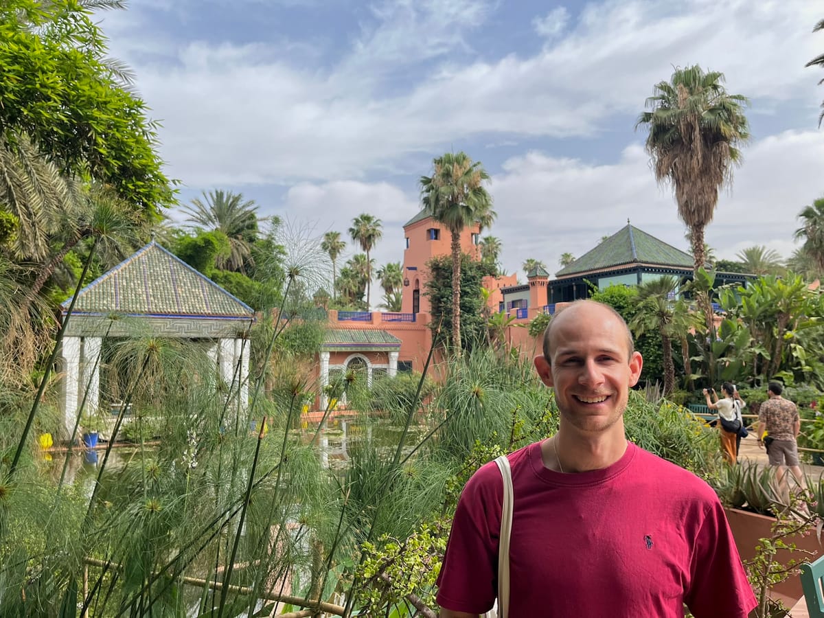 Smiling man in a maroon shirt standing in a lush, tropical garden with tiled pavilions and palm trees, under a partly cloudy sky