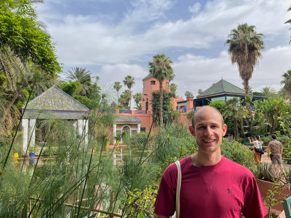 Smiling man in a maroon shirt standing in a lush, tropical garden with tiled pavilions and palm trees, under a partly cloudy sky